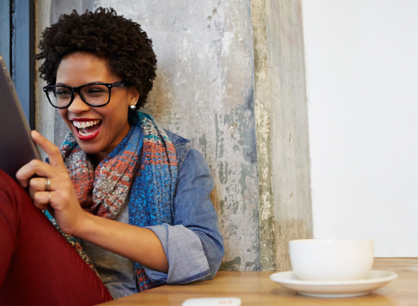 A lady sitting comfortably with her cup of coffee on a table and laughing and looking at her tablet